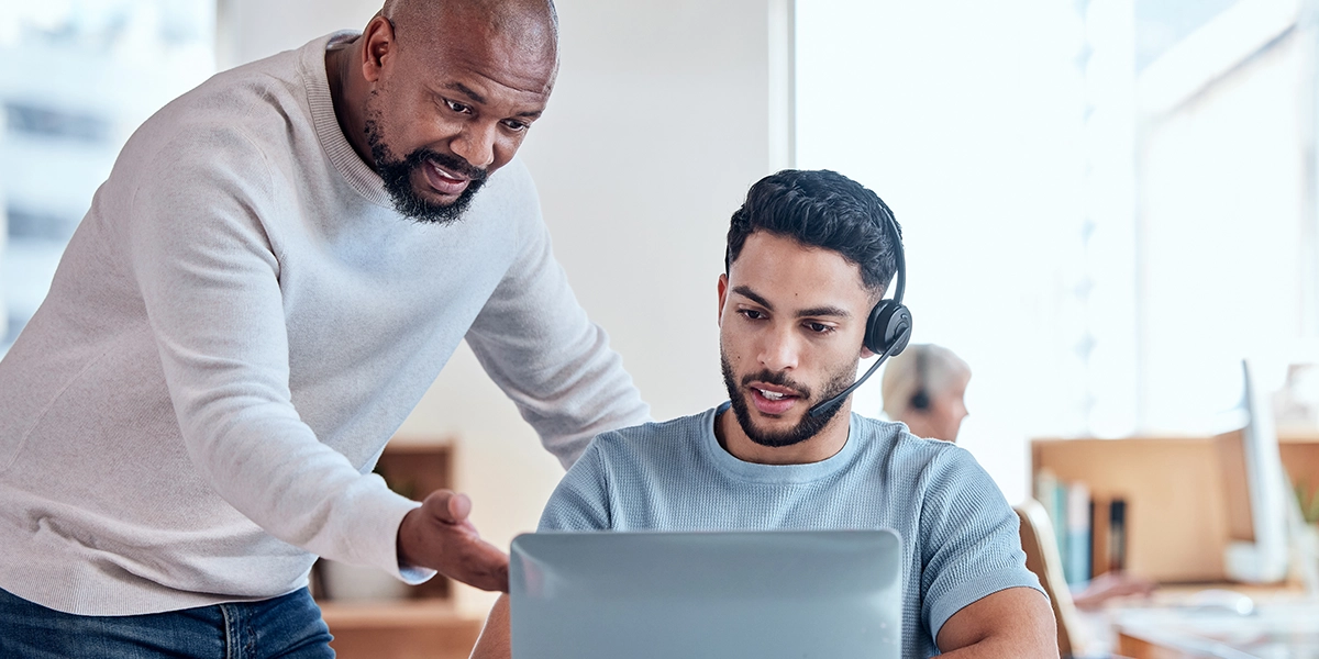 A imagem mostra um homem usando óculos de som e olhando para um laptops while outro homem olha. O laptops tem um fundo azul e o rosto do homem com óculos é escondido por os óculos. O homem na parte de trás está usando uma camisa branca e calças negras. A imagem está em foco e a iluminação é clara. A atmosfera é profissional e o cenário é uma sala de trabalho ou espaço de trabalho. A imagem é de boa qualidade e a resolução é alta. A imagem é adequada para uso em um contexto empresarial ou tecnológico.