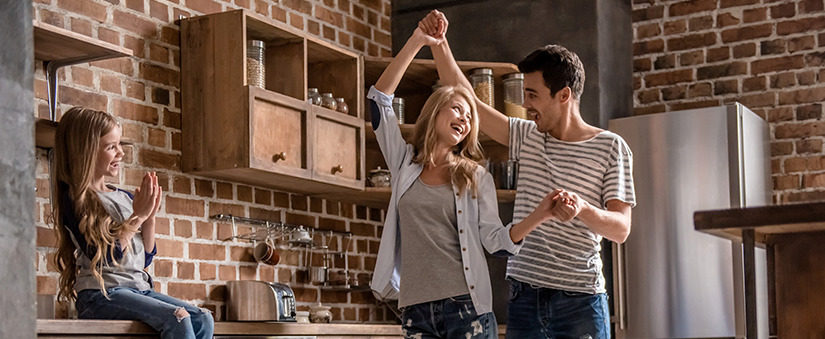 Casal dança na cozinha enquanto filha assiste sentada na bancada