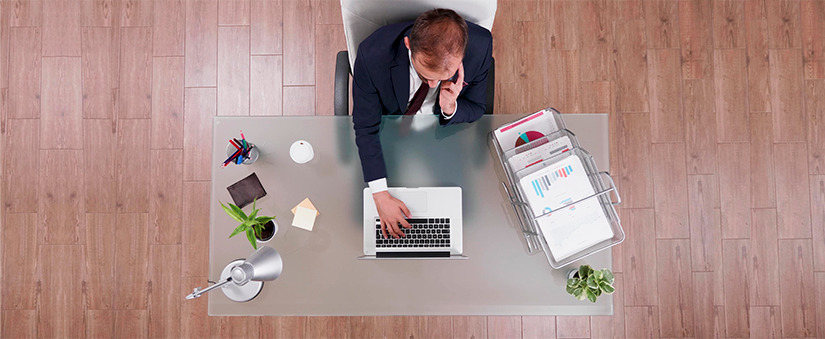 imagem de cima de um homem de terno sentado à frente de uma mesa com um notebook, papeis com gráficos e itens de escritório.