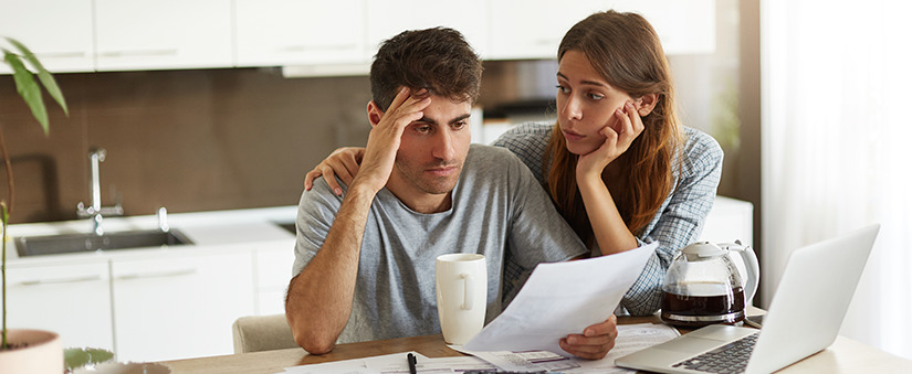 um homem segurando um papel, com uma das mãos na cabeça e uma mulher sentados à frente de uma mesa com um computador, com expressão de preocupação.