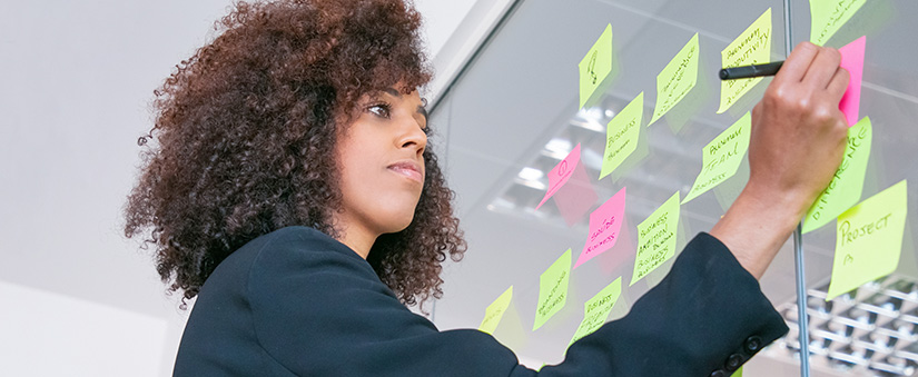 Ferramentas de Produtividade: mulher escrevendo em post it numa parede de vidro.