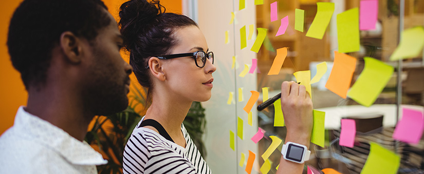 um homem e uma mulher em frente a uma parede de vidro com muitos postits colados.