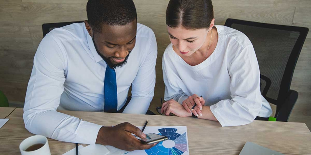 Técnico em edificações: duas pessoas sentadas à frente de uma mesa olhando para um celular e alguns papeis com gráficos.