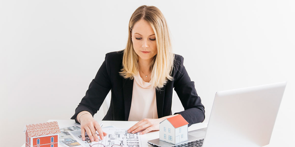 Site de imóveis: imagem de uma mulher sentada à frente de uma mesa com um notebook, alguns papeis e miniatura de casas de papel.