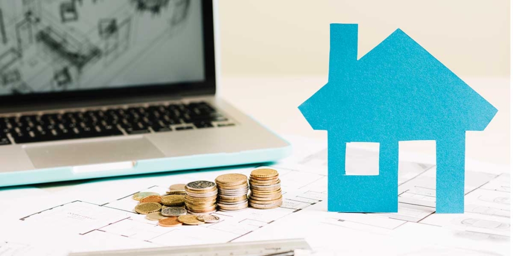 investir no mercado imobiliário: imagem de uma casa de papel ao lado de algumas moedas empilhadas. No fundo um notebook mostrando a planta de uma casa.