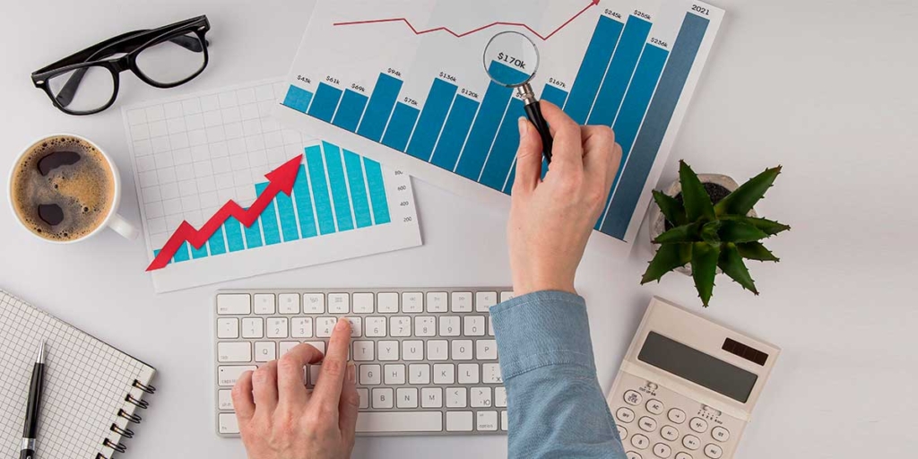 spin selling: imagem vista de cima de uma mesa com uma mão em cima de um teclado de computador, outra segurando uma lupa e apontando para papeis com gráficos.