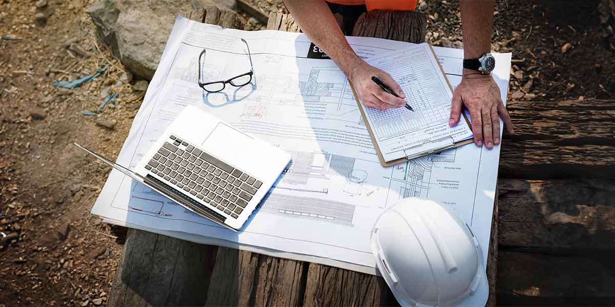 diário de obras: mesa vista de cima com uma planta de casa, um notebook, um capacete e mãos de uma pessoa escrevendo num papel.