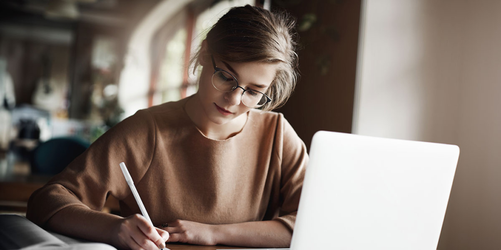 Script para corretor de imóveis: mulher escrevendo num papel em frente a um notebook.