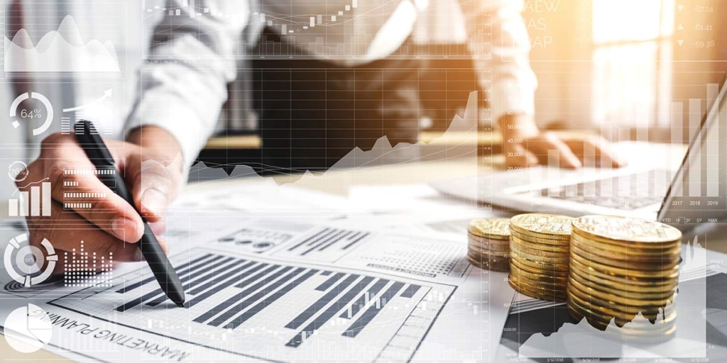 O que é lig investimento: close nas mãos de uma pessoa em cima de uma mesa cheia de papeis, moedas empilhadas e um notebook.