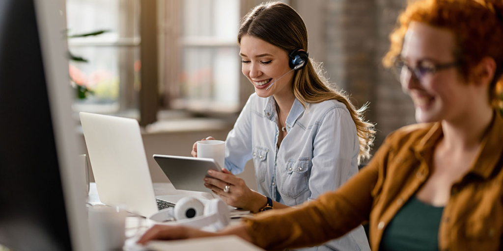 Atendimento ao cliente na construção civil: imagem de duas mulheres numa mesa de escritório com notebooks, headphones, etc.