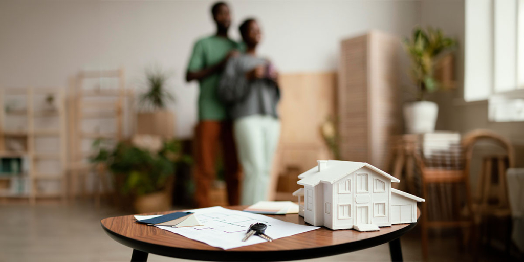 Casa paulista: imagem de uma mesa com uma casa em miniatura em cima, ao lado de uma chave. No fundo, em desfoque, duas pessoas olham para frente.