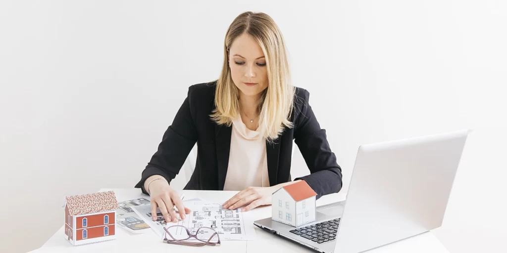 Gestor Imobiliário: imagem de uma mulher sentada em frente a uma mesa com muitos papeis, um óculos de grau, miniaturas de casa e um notebook.
