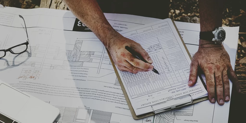 ABNT da Construção Civil: imagem de uma mesa vista de cima com uma planta de empreendimento. Acima do papel da planta, mãos de uma pessoa preenchendo uma tabela.