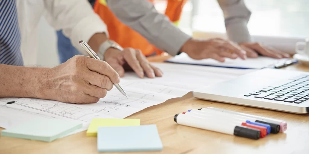 ABNT da Construção Civil: close nas mãos de pessoas numa mesa com muitos papeis, canetas e um notebook.