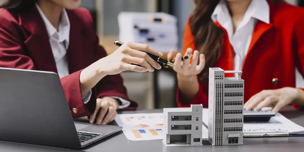 PropCo: foco nas mãos de duas mulheres gesticulando, com lápis na mão. Elas estão num escritório, na frente de uma mesa com papeis com gráficos, notebook e miniatura de edifícios.