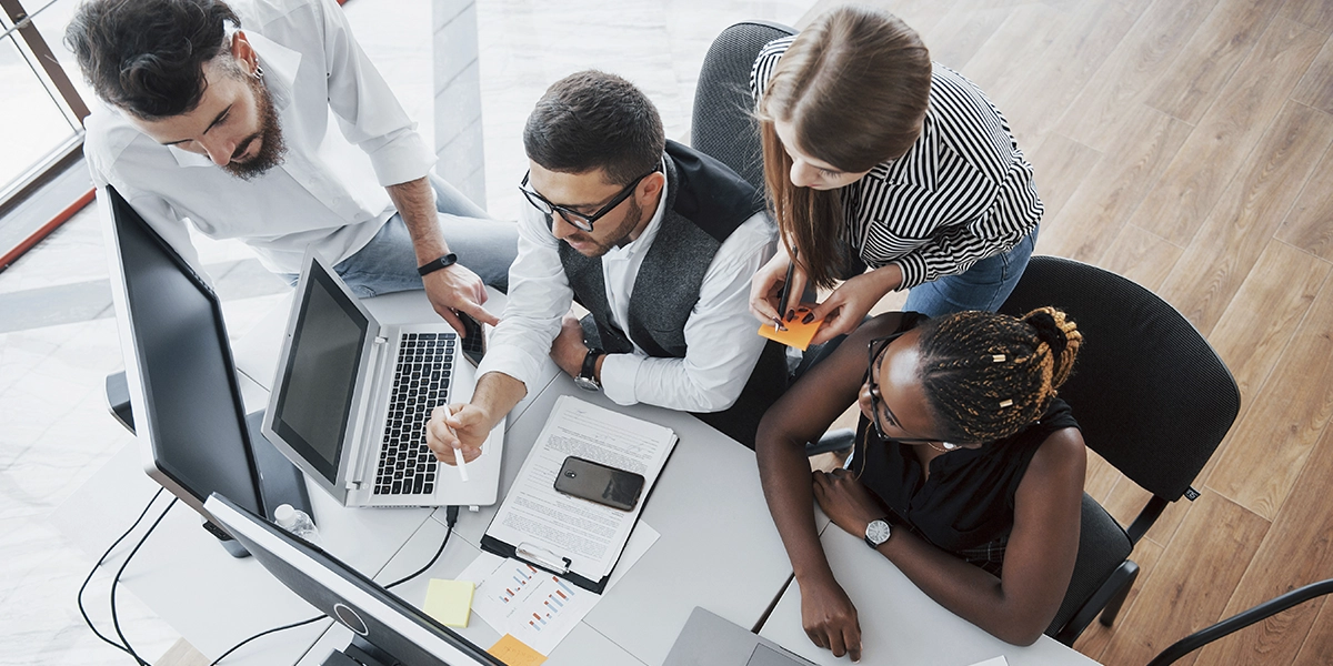 Administração de construtoras: imagem vista de cima de quatro pessoas num escritório, diante de uma mesa, olhando para um notebook