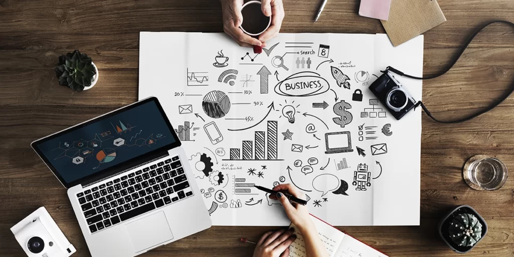 Inbound Marketing Imobiliário: imagem vista de cima de uma mesa com mãos de duas pessoas. Uma segura uma xícara de café, enquanto a outra aponta um lápis para um papel no centro da mesa com vários ícones de conexão, dinheiro, trabalho, alguns gráficos, balões de fala, setas, etc.
