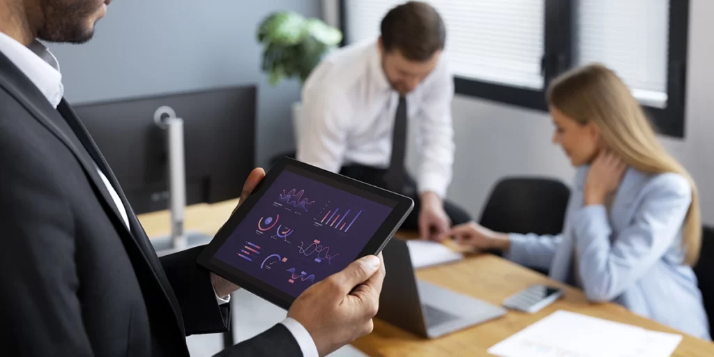 Portifólios imobiliários: imagem de um homem num escritório segurando um tablet com um dashboard na tela. Em segundo plano um homem e uma mulher conversam.