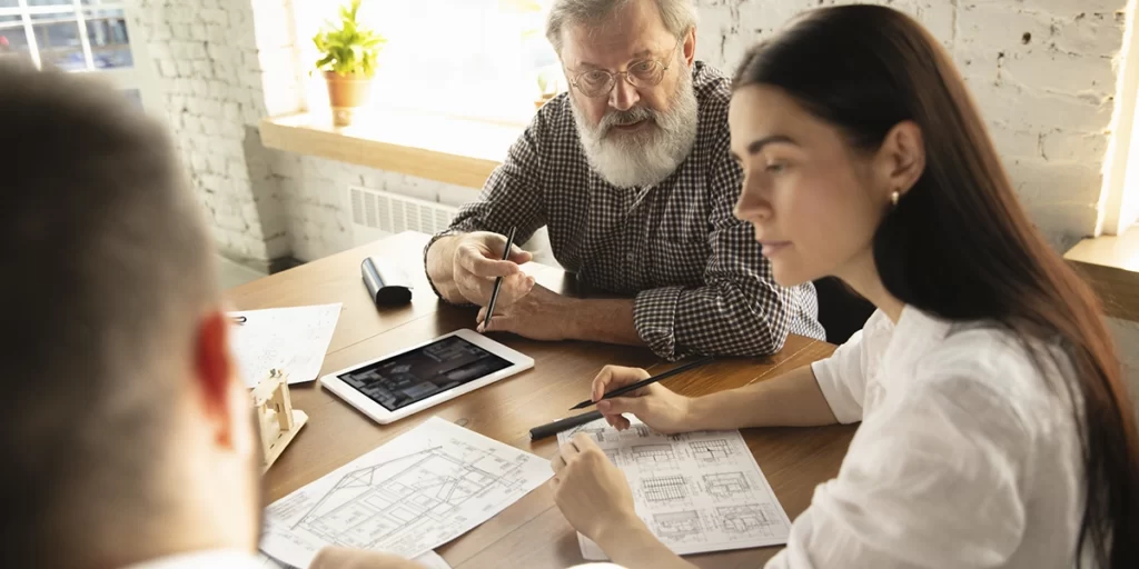 Administração de Imóveis: imagem de três pessoas conversando diante de uma mesa. Em cima da mesa vemos alguns papeis de planta de imóveis e um tablet.