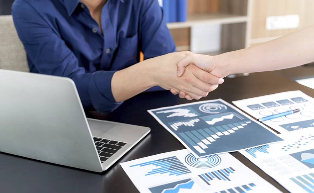 Como prospectar mais clientes: close num aperto de mãos entre duas pessoas de lados opostos de uma mesa. Na mesa um notebook e alguns papeis com gráficos coloridos.