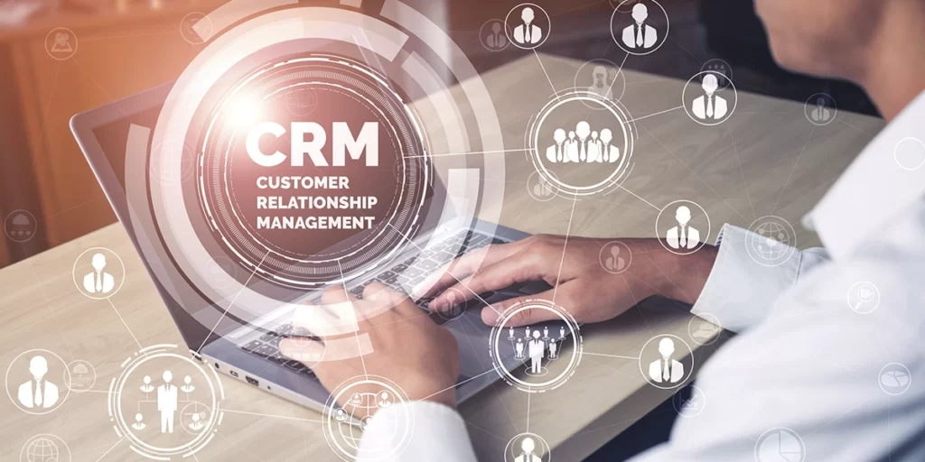 Software Imobiliário: close nas mãos de uma pessoa digitando num notebook. Em destaque na imagem, a sigla "CRM" e algumas ilustrações de pessoas em volta.