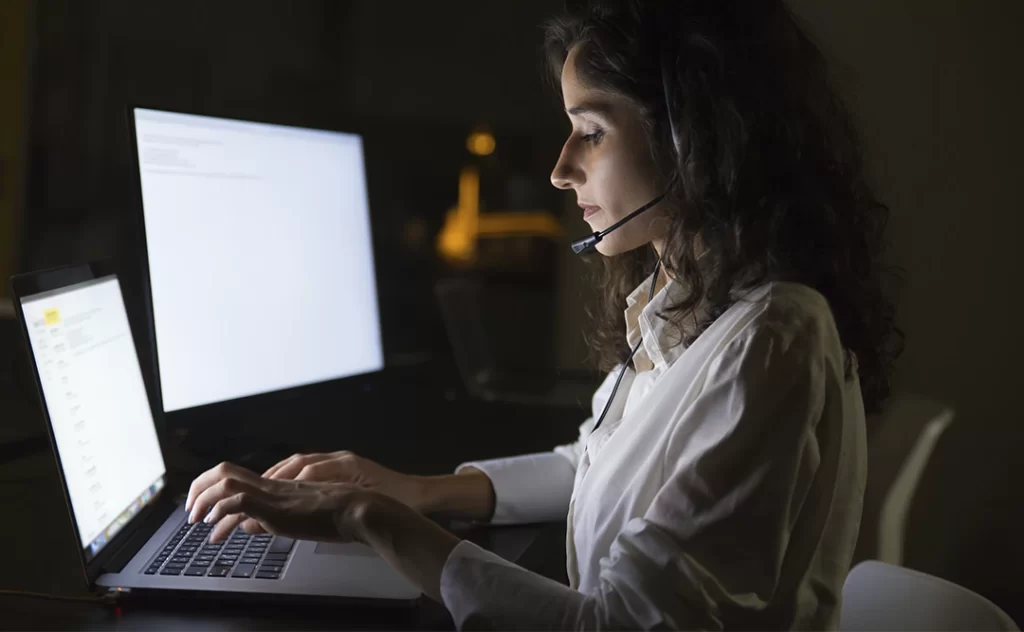  vantagens da inteligência artificial: imagem de uma mulher sentada à frente de um computador numa sala com poucas luzes, destacando as luzes das telas na mesa. Ela sua um fone com microfone.