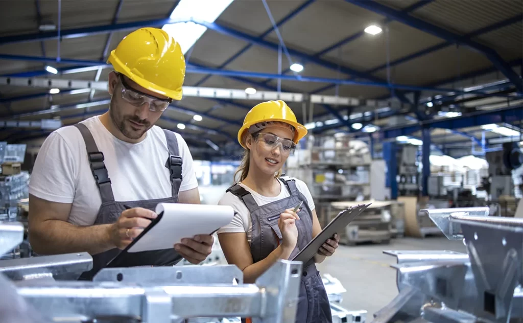 Gestão da qualidade na construção civil: A imagem mostra dois indivíduos em um ambiente industrial. Ambos estão usando capacetes amarelos e vestindo trajes de proteção. O indivíduo à esquerda está segurando um caderno aberto e parece estar examinando algo escrito nele. O indivíduo à direita está segurando um tablet e parece estar trabalhando ou consultando informações. 