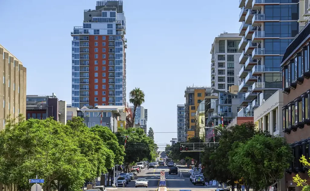 Indicadores imobiliários: A imagem mostra uma vista de uma rua em um ambiente urbano. Há edifícios altos ao longo da borda da rua, incluindo prédios residenciais e possivelmente comerciais. A rua está bem iluminada e parece ser um dia claro. Há muitos carros estacionados e em movimento, e a paisagem é acentrada pela presença de árvores ao longo da borda da rua, o que dá um contraste verde à cena urbana. A rua é ampla e parece ser uma via principal, com sinais de trânsito e possivelmente uma estrutura de transporte aéreo ou ferroviário ao fundo.