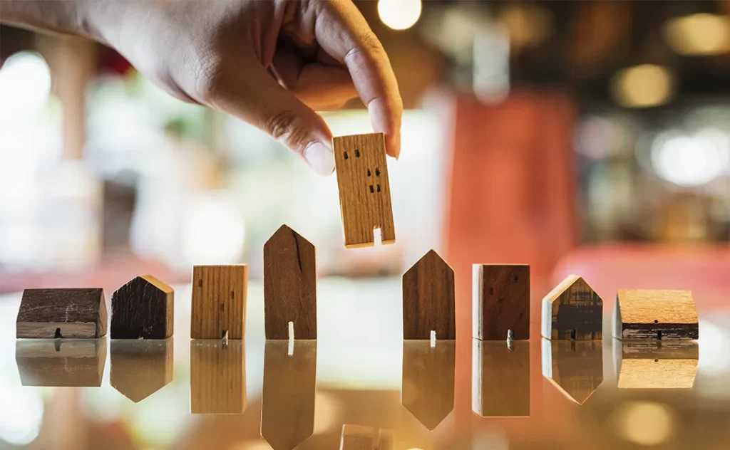Indicadores imobiliários: A imagem mostra uma mão humana segurando um pequeno modelo de casa feito de madeira. Há outros modelos de casas de madeira de diferentes tamanhos e estilos dispostos em uma superfície, possivelmente um tábano ou uma superfície de madeira, com suas reflexões visíveis na superfície. A foto parece ter sido tirada em um ambiente interno com iluminação artificial, e a mão está na parte superior da imagem, destacando a interação com o modelo de casa.