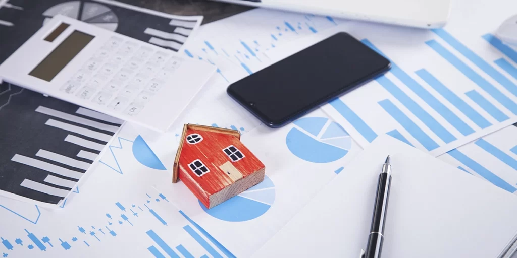 Índice Imobiliário: A imagem mostra uma superfície de trabalho com vários itens relacionados a negócios e finanças. Há um aparelho de cálculo, um celular, um lápis, um pequeno modelo de casa, um caderno e algumas folhas de papel com gráficos e dados. A disposição dos itens sugere um ambiente de trabalho ou estudo financeiro ou de negócios.