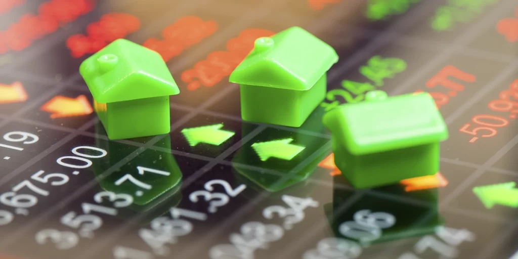 Índice Imobiliário: A imagem mostra três peças verdes de um brinquedo, que parecem ser peças de uma casa, sobre um fundo com gráficos financeiros. O fundo apresenta gráficos de desempenho de ações ou índices financeiros, com ícones de setas vermelhas e verdes, indicando movimentos de preço. As casas em miniatura são representação visual simbólica do mercado financeiro, possivelmente sugerindo investimentos em imóveis ou ações de empresas relacionadas ao setor imobiliário.