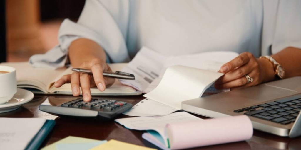 Pagamento de comissões: imagem das mãos de uma pessoa sentada à frente de uma mesa com muitos papeis, uma calculadora, um notebook e xícara de café.