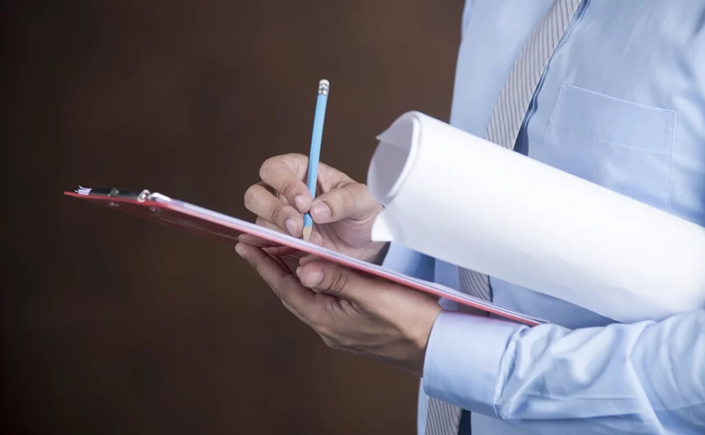 Anotação de Responsabilidade Técnica: A imagem mostra uma mão de um indivíduo vestindo um terno azul escrevendo com uma caneta azul sobre um caderno vermelho. O caderno está aberto e parece estar sendo usado para escrever ou tomar notas. O fundo é desfocado, mas parece ser um ambiente interno com uma parede de cor escura.