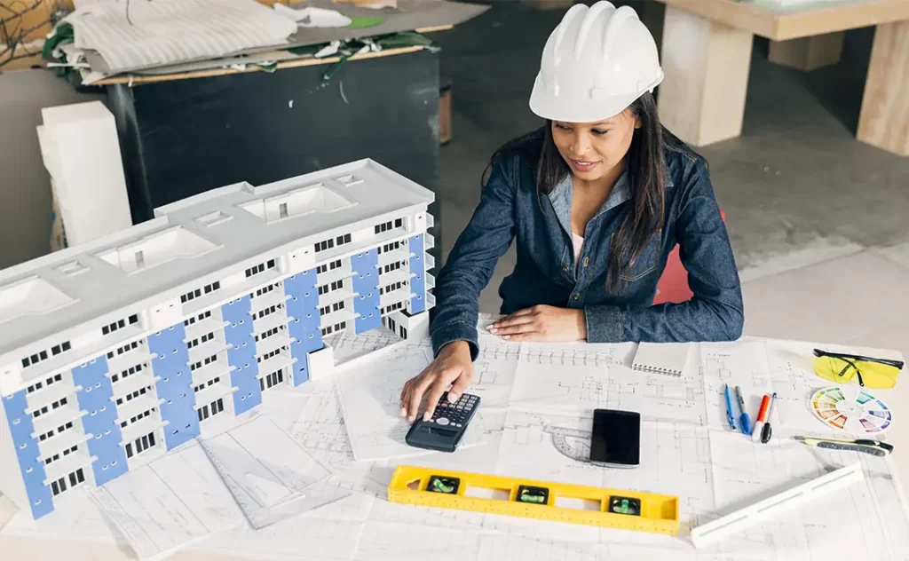 Anotação de Responsabilidade Técnica: A imagem mostra uma mulher sentada ao redor de um tabela com planos arquitetônicos e material de construção. Ela está usando um capacete branco e veste uma jaqueta azul. A mulher parece estar trabalhando ou estudando os planos, possivelmente em um ambiente de construção ou projeto arquitetônico. Há uma estrutura de construção em miniatura ao lado dela, sugerindo que ela pode estar trabalhando em um projeto de arquitetura ou engenharia.