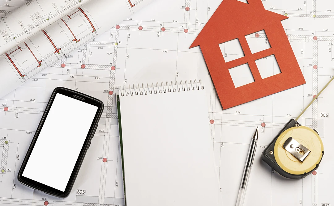 A imagem mostra uma variedade de itens relacionados a projetos de construção ou arquitetura dispostos sobre planos de construção. Há um smartphone, um caderno aberto, um lápis, um compasso, um modelo de casa em papel e alguns planos de construção abertos. Tudo isso sugere que alguém está trabalhando em um projeto de arquitetura ou engenharia.