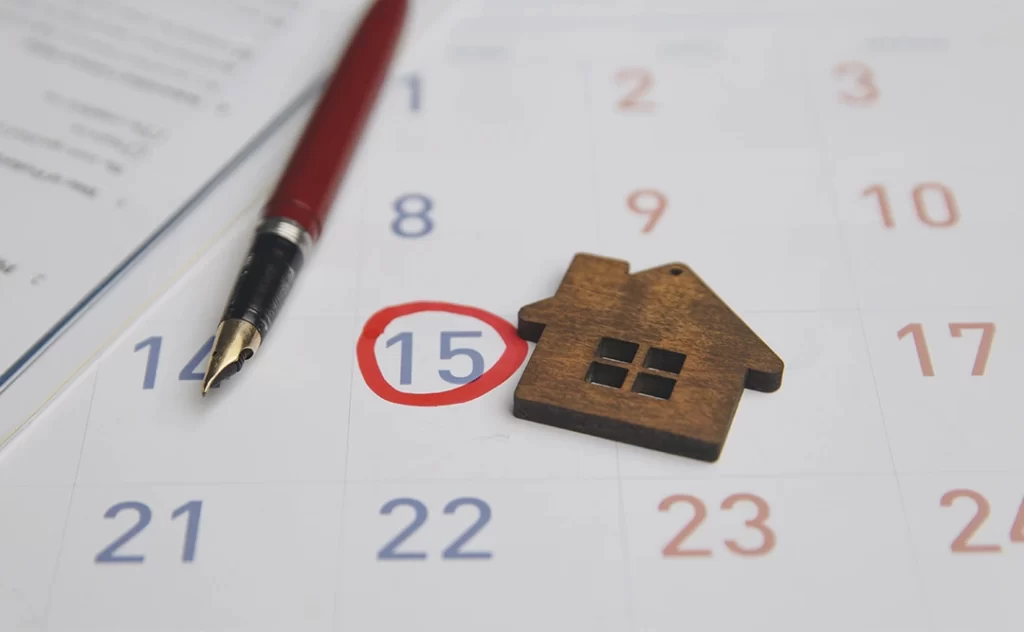 Calendário imobiliário: A imagem mostra um calendário de mês com o dia 15 marcado com um círculo vermelho. Ao lado do calendário, há um ponto de escrita preto e um pequeno logotipo de uma casa, que parece ser um marcador de calendário ou um adesivo decorativo. O calendário está sobre um papel com texto, e a data marcada pode indicar um evento importante, como uma data de mudança ou uma data de contrato de aluguel.