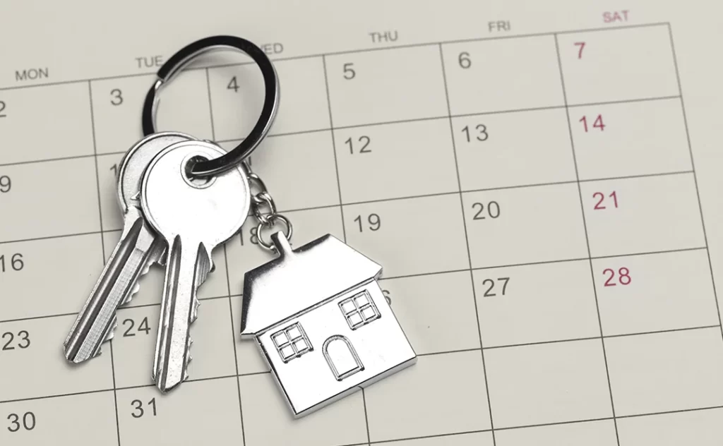 Calendário imobiliário: A imagem mostra uma chave de porta com um anel de segurança e um pequeno logotipo de uma casa, colocada sobre um calendário de mês. O calendário está em um papel ou material de cor clara, e as datas estão escritas em preto. A chave e o anel de segurança são de um material que parece ser metal, possivelmente prateado ou dourado, e estão em contato com o calendário. A imagem tem um estilo simples e é focada na chave e no calendário, sugerindo uma conexão entre a segurança de uma propriedade e o controle do tempo ou eventos.