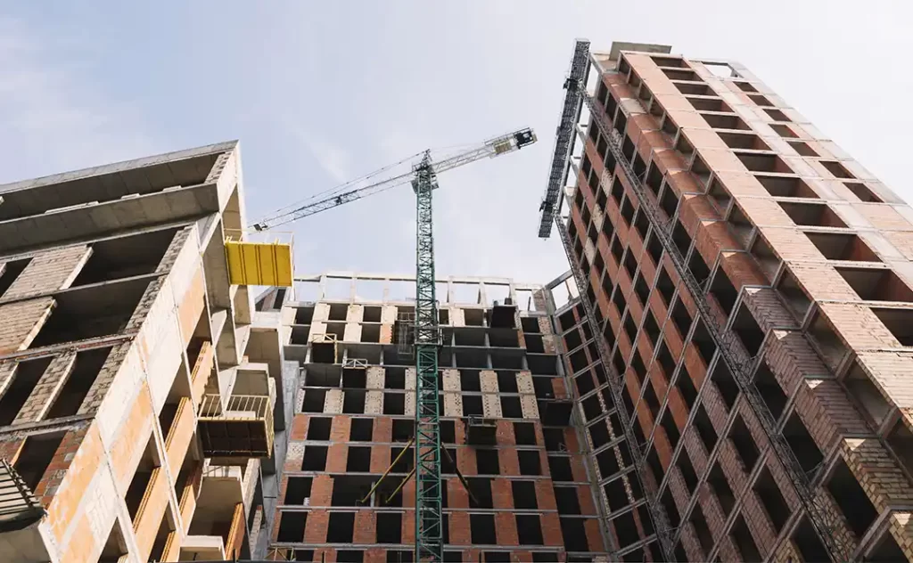 Como calcular o bdi: A imagem mostra um edifício em construção em um ambiente urbano. Há um craneo de construção ao lado do edifício, indicando que o projeto ainda está em andamento. O edifício apresenta várias camadas de estrutura, com algumas partes ainda sem paredes, o que sugere que a construção está em um estágio avançado. O céu está claro, sugerindo um dia ensolarado.