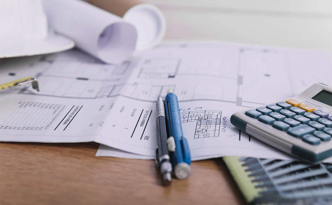 Como calcular o bdi: A imagem mostra uma superfície com vários itens relacionados a trabalho de contabilidade ou finanças. Há um caderno aberto com páginas de contabilidade, um calcador de cálculo, um lápis e um caneta. Também há um chapéu de construtor e um rolo de papel, sugerindo que o trabalho pode estar relacionado a projetos de construção ou arquitetura. A cena parece estar em um ambiente de trabalho ou escritório.