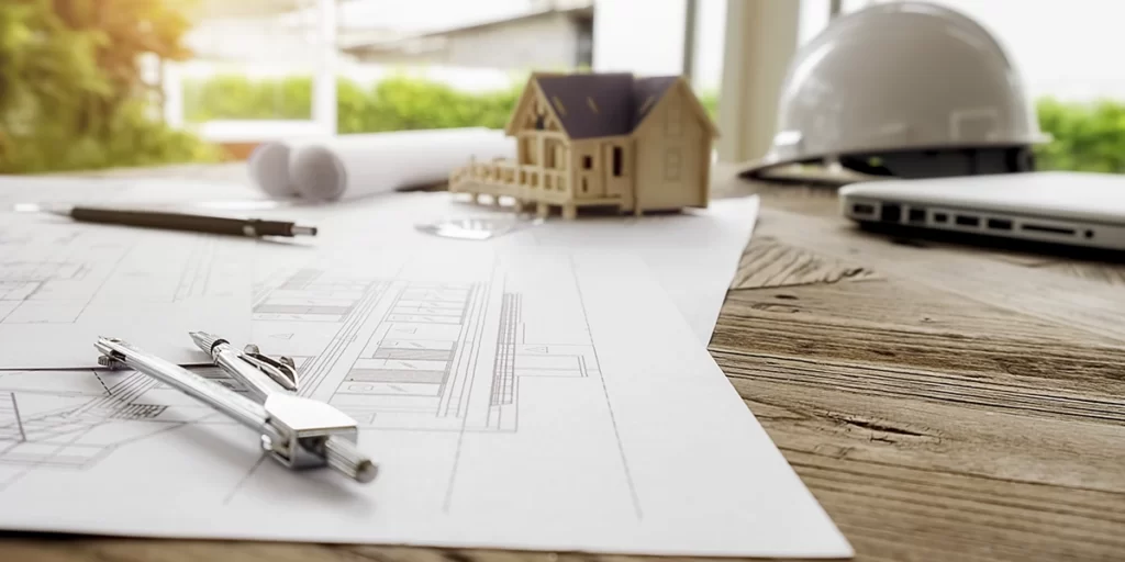 Documentação de terreno: imagem de uma mesa com alguns papeis de planta de imóvel, um transferidor, um lápis. Ao fundo uma casa em miniatura, um capacete de obra e um notebook.