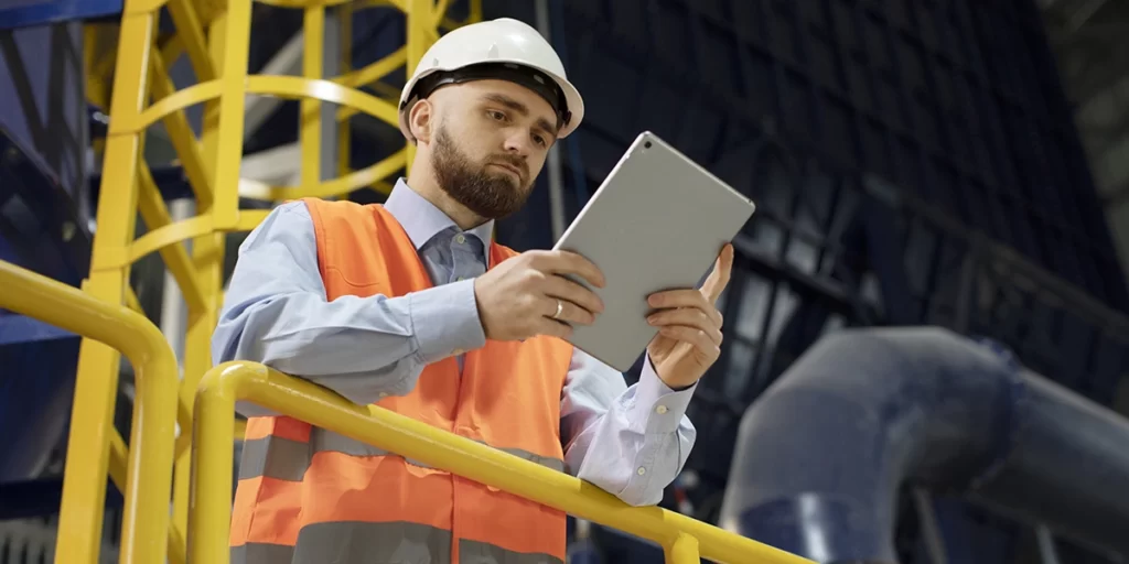 Gestão de mão-de-obra: A imagem mostra um homem vestindo um capacete de segurança branco e um jaqueta de segurança laranja com linhas pretas, que é comum em ambientes industriais. Ele está segurando um tablet e parece estar olhando para ele, possivelmente ver informações ou trabalhando. O homem tem uma barba curta e está usando uma camisa azul. Ao fundo, há uma estrutura industrial com tubos e partes de metal, sugerindo que o ambiente é um local de trabalho industrial.