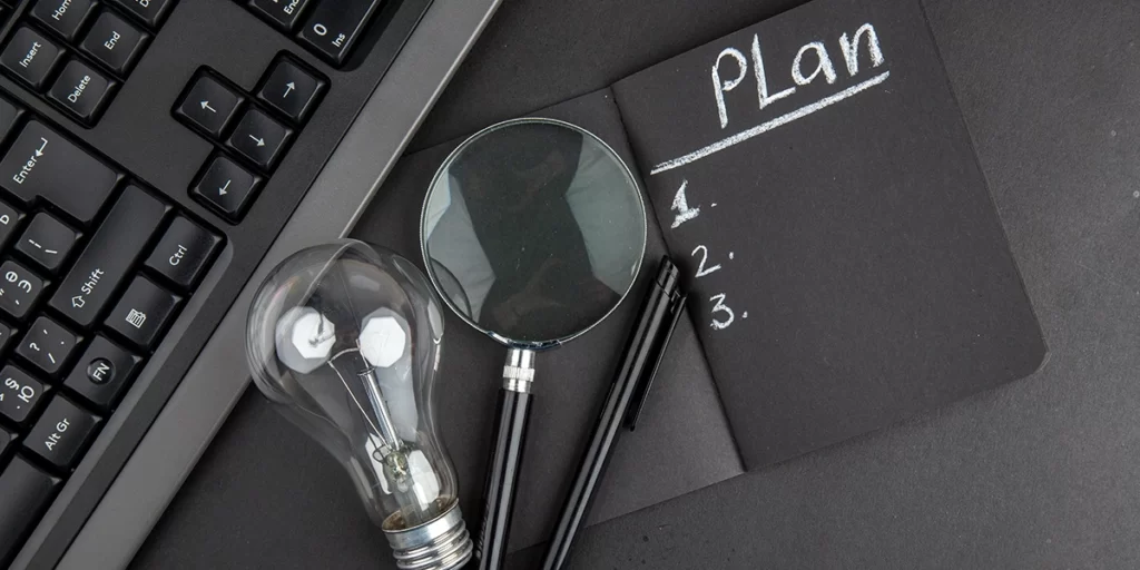 Plano de Negócio: A imagem mostra uma escrivaninha preta com o texto "Plan" escrito em branco no topo. Abaixo do texto, há três números: "1.", "2." e "3.", também escritos em branco. Ao lado da escrivaninha, há um teclado preto com alguns botões visíveis. Ao lado do teclado, há um par de óculos de armação preta e lentes brancas, e um par de lâmpadas de armação preta e vidro claro. A lâmpada está parcialmente dentro da escrivaninha, e o par de óculos está parcialmente dentro da lâmpada. A imagem tem um estilo moderno e parece ser uma representação visual de planejamento ou ideias.
