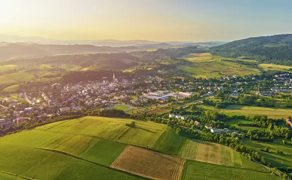 Terrenista: A imagem mostra uma paisagem rural com uma vista aérea. Há uma cidade com edifícios de arquitetura tradicional, rodeada por campos de grama verde e áreas de cultivo. Ao fundo, há uma série de colinas e montanhas, sugerindo uma área de terra baixa ou uma região costeira. O sol está baixo no horizonte, indicando que pode ser perto do horário do pôr do sol ou do nascer do sol, o que dá à cena um aspecto tranquilo e pitoresco.