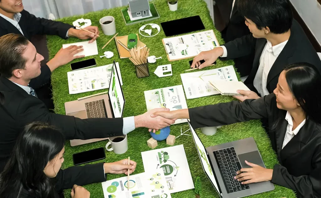 Terrenista: A imagem mostra uma cena de negociações ou reunião de negócios em um ambiente moderno e casual. Há cinco pessoas sentadas em volta de uma mesa com grama artificial verde, sugerindo um ambiente de trabalho eficiente e ambiente. Dois homens estão se dando um abraço de mão, indicando um acordo ou um momento de celebração. As outras pessoas estão engrossadas em documentos e dispositivos, como laptops e celulares, sugerindo que estão trabalhando ou discutindo negócios. A mesa está bem organizada com notas, apresentações e outros materiais relacionados ao trabalho. A atmosfera é profissional, mas com um toque de casualidade, possivelmente indicando uma reunião de negócios mais informal ou uma equipe que trabalha de forma colaborativa e descontraída.