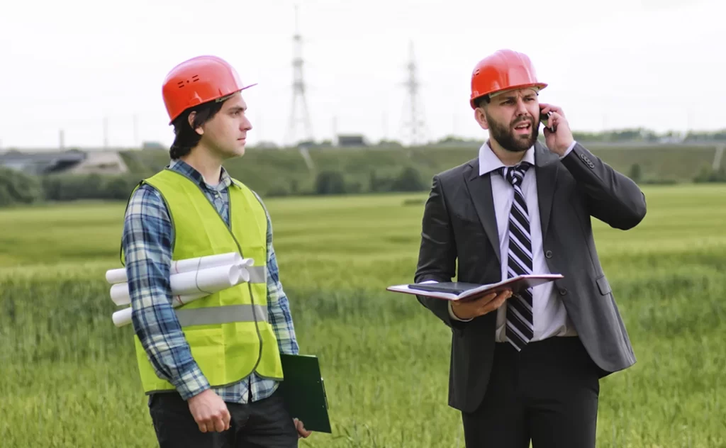 Terrenista: A imagem mostra dois homens em um ambiente ao ar livre, possivelmente um campo ou uma área de construção. O homem à esquerda está vestindo uma jaqueta amarilla de segurança, um capacete vermelho e uma camisa de manga curta com padrões. Ele parece estar olhando para o lado, possivelmente atento a algo fora da cena. O homem à direita está usando um terno preto, uma camisa branca com uma gravata preta e branca, e também um capacete vermelho. Ele está falando em um telefone celular e parece estar segurando algum material, talvez um livro ou um folheto. Ambos parecem estar trabalhando juntos em um projeto ou uma tarefa.