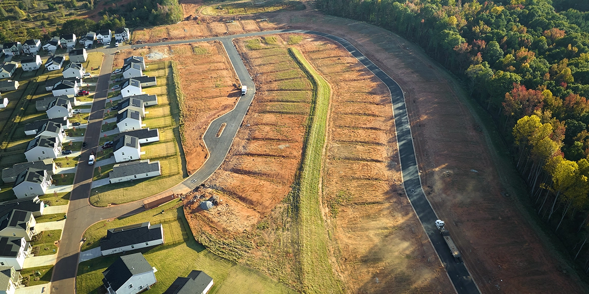 compra de lotes: Imagem vista de cima de uma vizinhança residencial com casas ao longo de uma estrada. As casas são feitas de madeira e têm grandes janelas e portas. Existem árvores em cada lado da estrada e o céu é claro e azul