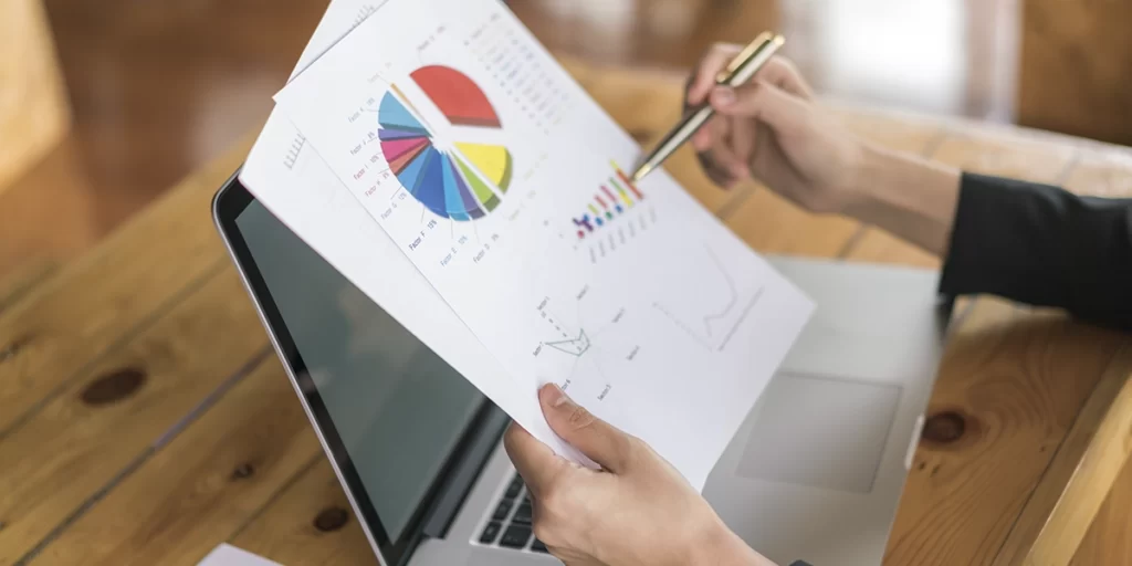 diagrama de ishikawa: A imagem mostra uma mão de um indivíduo sentado em frente a um laptop aberto. A mão está segurando uma folha de papel com gráficos e dados, possivelmente um relatório ou um estudo estatístico. A outra mão está segurando um caneta, sugerindo que o indivíduo pode estar anotando ou estudando os dados na folha. O ambiente parece ser um espaço de trabalho ou escritório, com uma mesa de madeira e uma cadeira ao fundo.