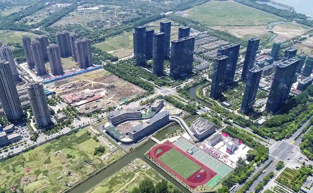 Urbanização e construção civil: A imagem é uma foto aérea de uma área urbana. Há uma variedade de edifícios altos, incluindo prédios residenciais e possivelmente comerciais ou oficiais, que são dispostos em um padrão regular. Há também áreas de terra nua e campos abertos, além de uma estrutura que parece ser um complexo de atividades físicas ou esportivas, com campos de futebol e outros espaços de jogo. Há uma corrente de água que atravessa a área, e a vegetação é predominantemente verde, sugerindo que há parques ou áreas de florestação. A imagem mostra uma mistura de construções e espaços abertos, indicando um desenvolvimento urbano que tenta equilibrar a infraestrutura com áreas verdes e espaços de lazer.