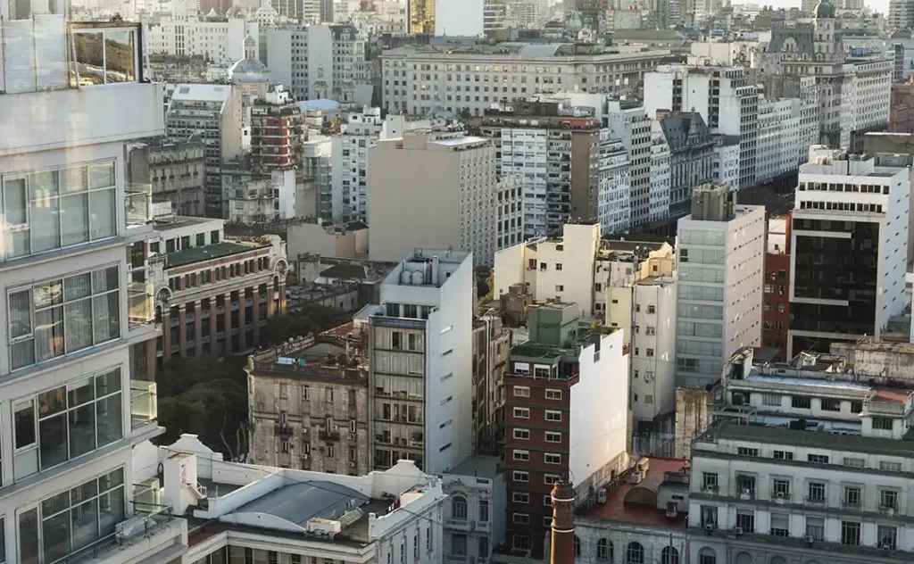 Urbanização e construção civil: A imagem mostra uma vista aérea de uma área urbana densa. Há uma variedade de edifícios de arquitetura moderna e histórica, com vários andares e cores que variam de brancos e claros a tons mais escuros. A paisagem é dominada por edifícios residenciais e comerciais, com alguns espaços abertos que parecem ser parques ou áreas de terra nua. A cena transmite uma sensação de grandeza e complexidade, com a cidade se estendendo até o horizonte.