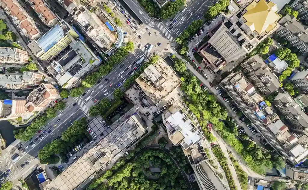 Urbanização e construção civil: A imagem é uma foto aérea de uma área urbana. Há uma estrutura arquitetônica complexa com edifícios de arranjo irregular, algumas de grande porte e outras de tamanho menor. Há uma estrada principal que se cruza com outra, formando um "X", e outras ruas que se bifurcam ou se cruzam. A paisagem é intercalada com áreas de vegetação, possivelmente parques ou jardins, que contrastam com o materialidade das construções. A imagem captura a densidade e a complexidade de uma cidade, com a interação entre arquitetura e espaço público.